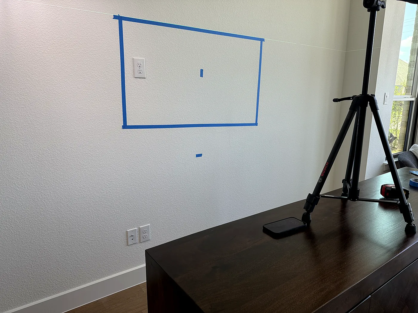Blue painter's tape outline marking the TV position on a wall beside a power outlet, with a laser level on a tripod